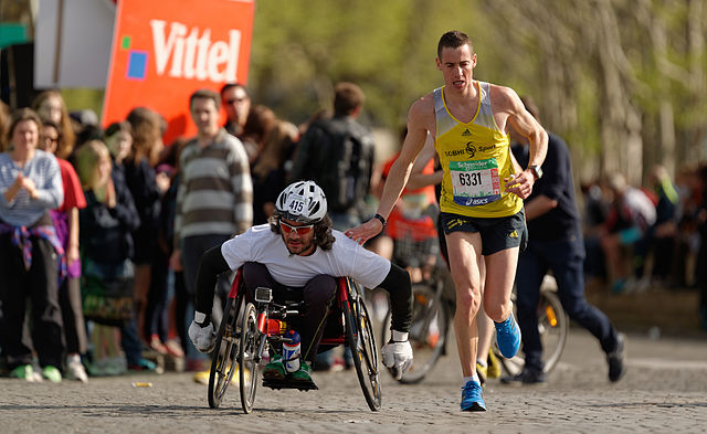 A man in a wheelchair and a man not in a wheel chair race together side by side 